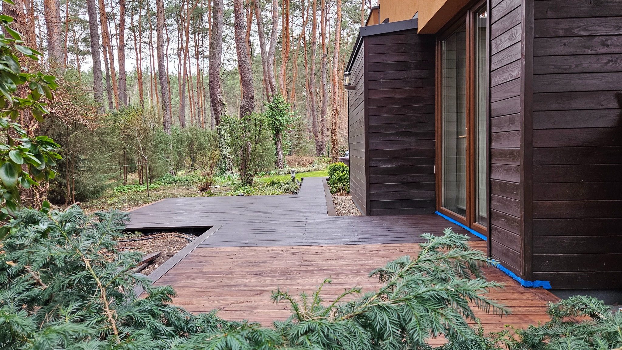 Ciemna, drewniana elewacja domu z tarasem w lesie. Część tarasu pomalowana, reszta w surowym drewnie. Widoczne zabezpieczenia okien. Praca w toku.