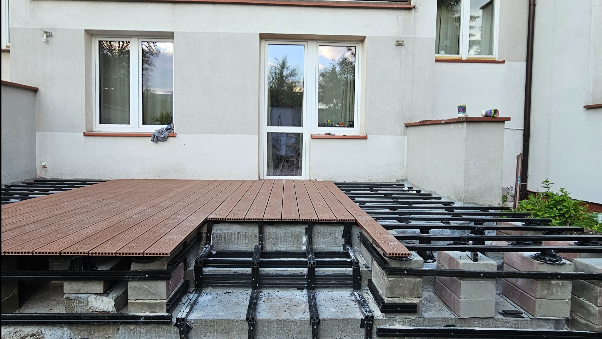 Montaż tarasu z deski kompozytowej na betonowej konstrukcji. Widoczne elementy mocujące i fragment ułożonej podłogi tarasu. Tło stanowi elewacja budynku z oknami.
