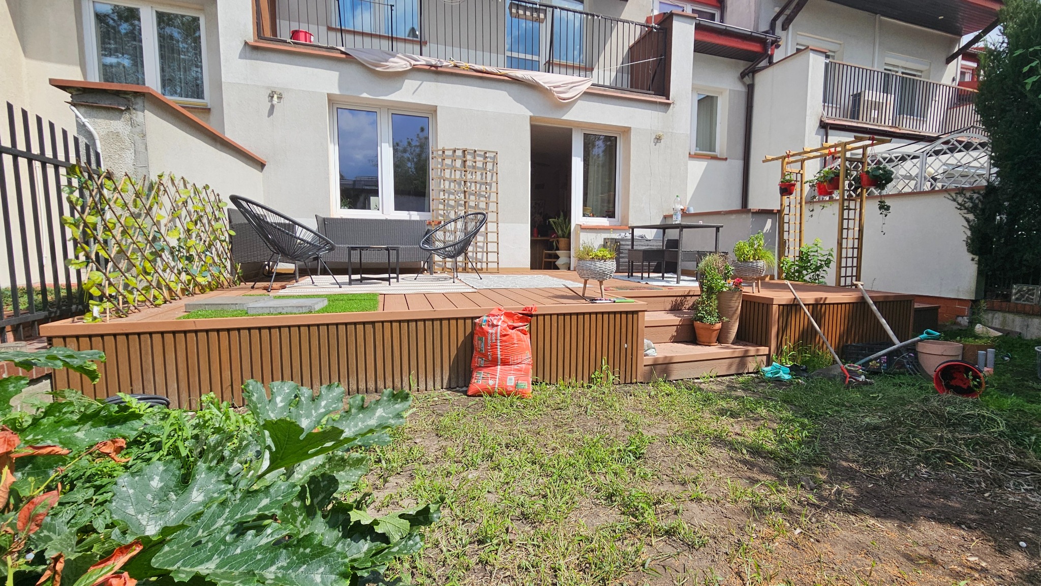 Drewniany taras z meblami ogrodowymi i pergolą w Warszawie. Widoczny worek z ziemią i narzędzia ogrodnicze. Ujęcie z poziomu trawnika, w tle budynek.