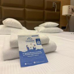 Góralskie Sprzątanie - Stos białych ręczników i ulotka firmy sprzątającej apartamenty 'Góralskie Sprzątanie' na łóżku w pokoju hotelowym. Ulotka reklamuje sprzątanie 5-gwiazdkowe.