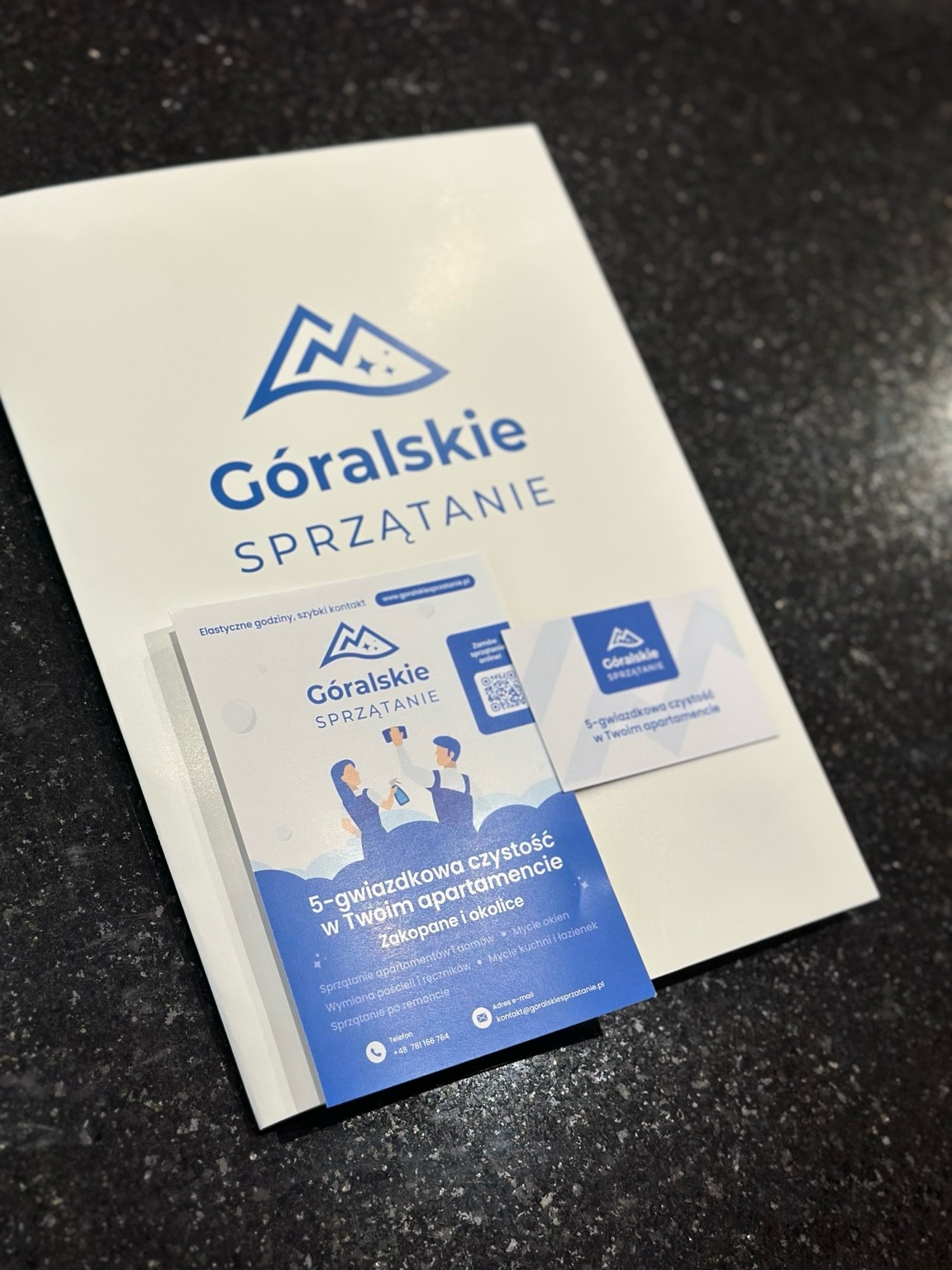 Materiały reklamowe firmy sprzątającej z Zakopanego. Ulotka i wizytówki z logo 'Góralskie Sprzątanie' na ciemnym blacie. Oferta 5-gwiazdkowej czystości w apartamentach.