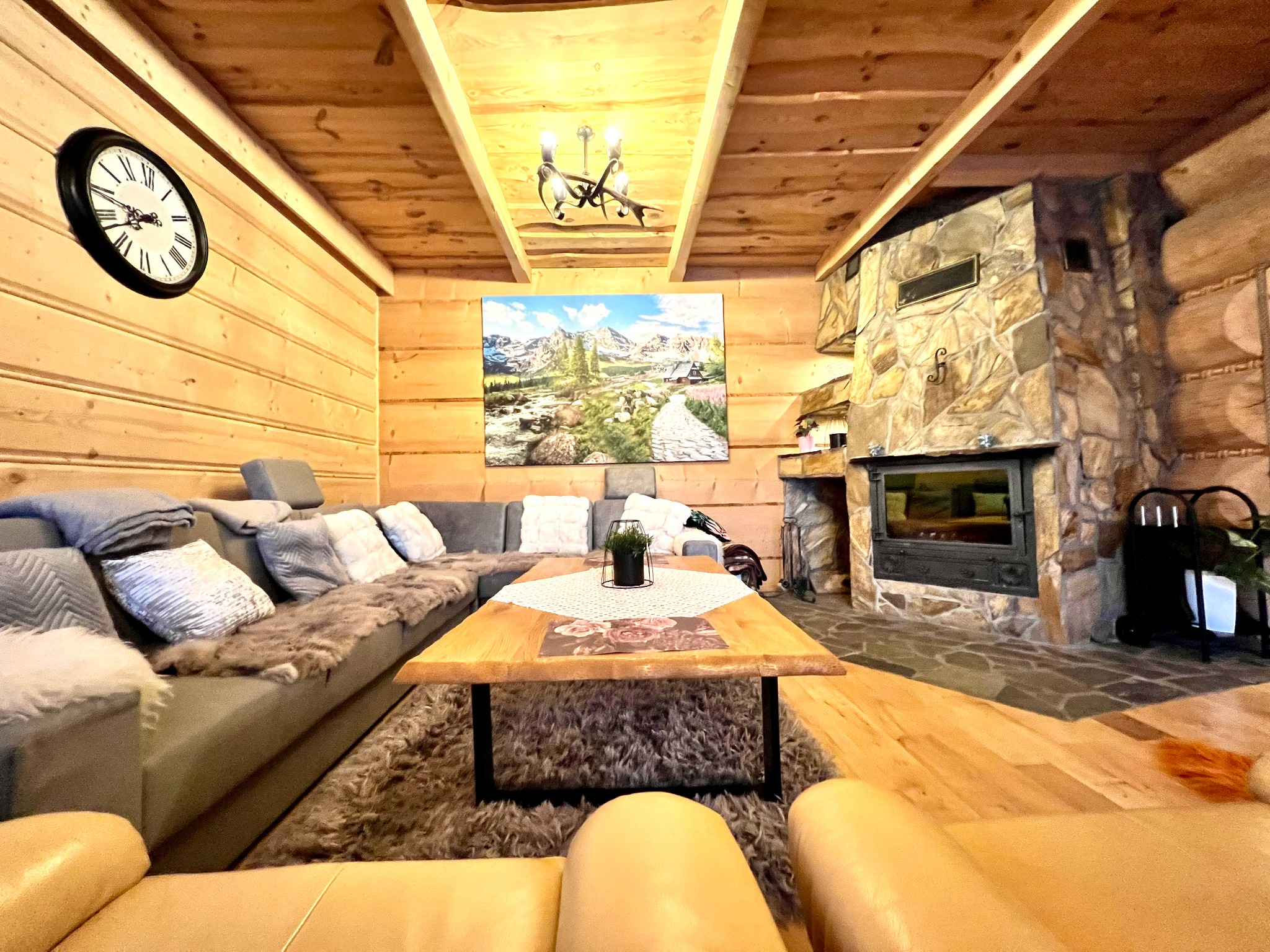 Rustykalny salon z drewnianymi ścianami, kamiennym kominkiem i dużym, narożnym kanapą. Na ścianie zegar i obraz z górskim pejzażem. Widok z perspektywy fotela.