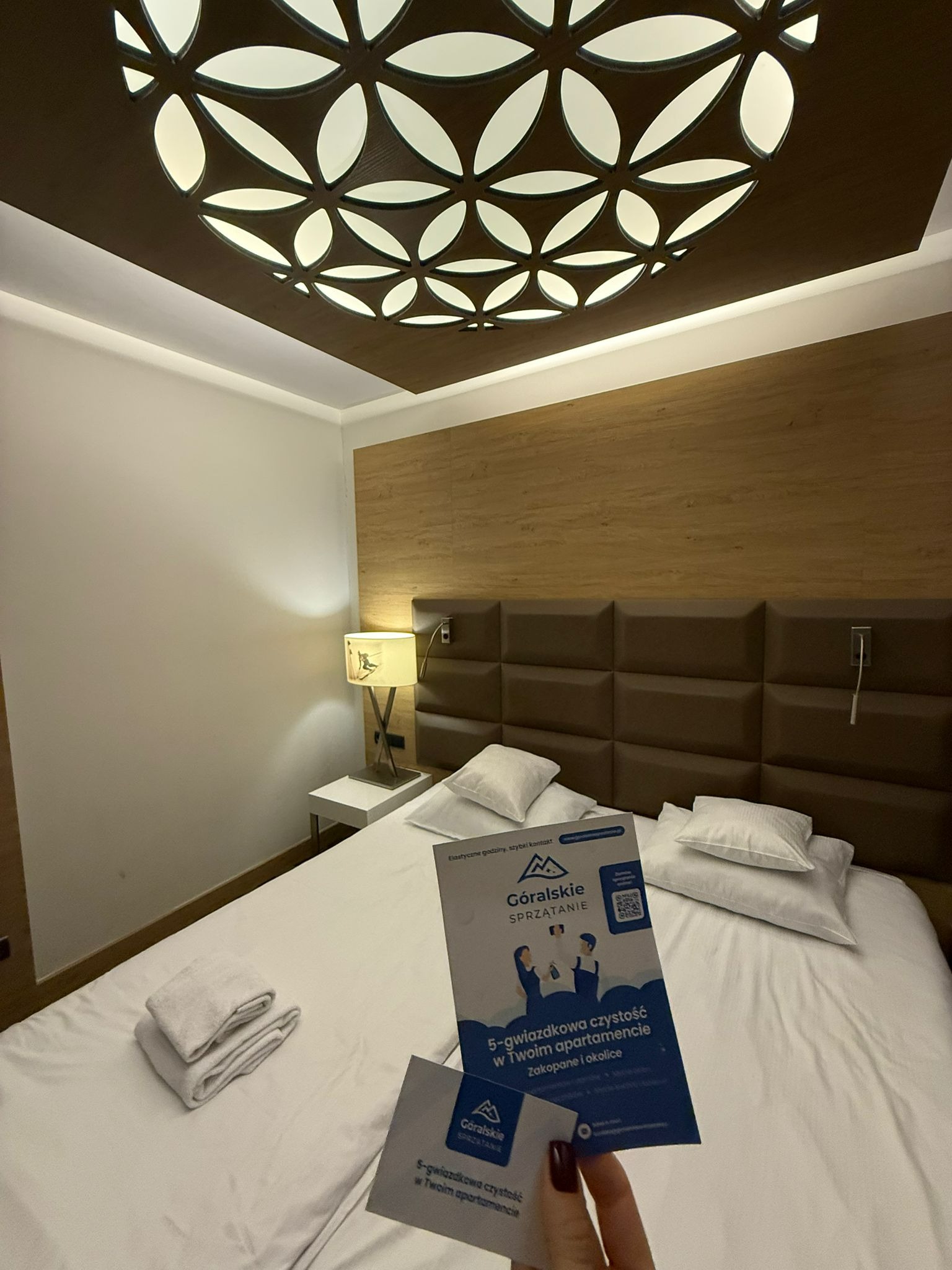 Czysty pokój hotelowy z broszurami 'Góralskie Sprzątanie' na łóżku. Dekoracyjna lampa sufitowa i starannie złożone ręczniki podkreślają dbałość o detale.