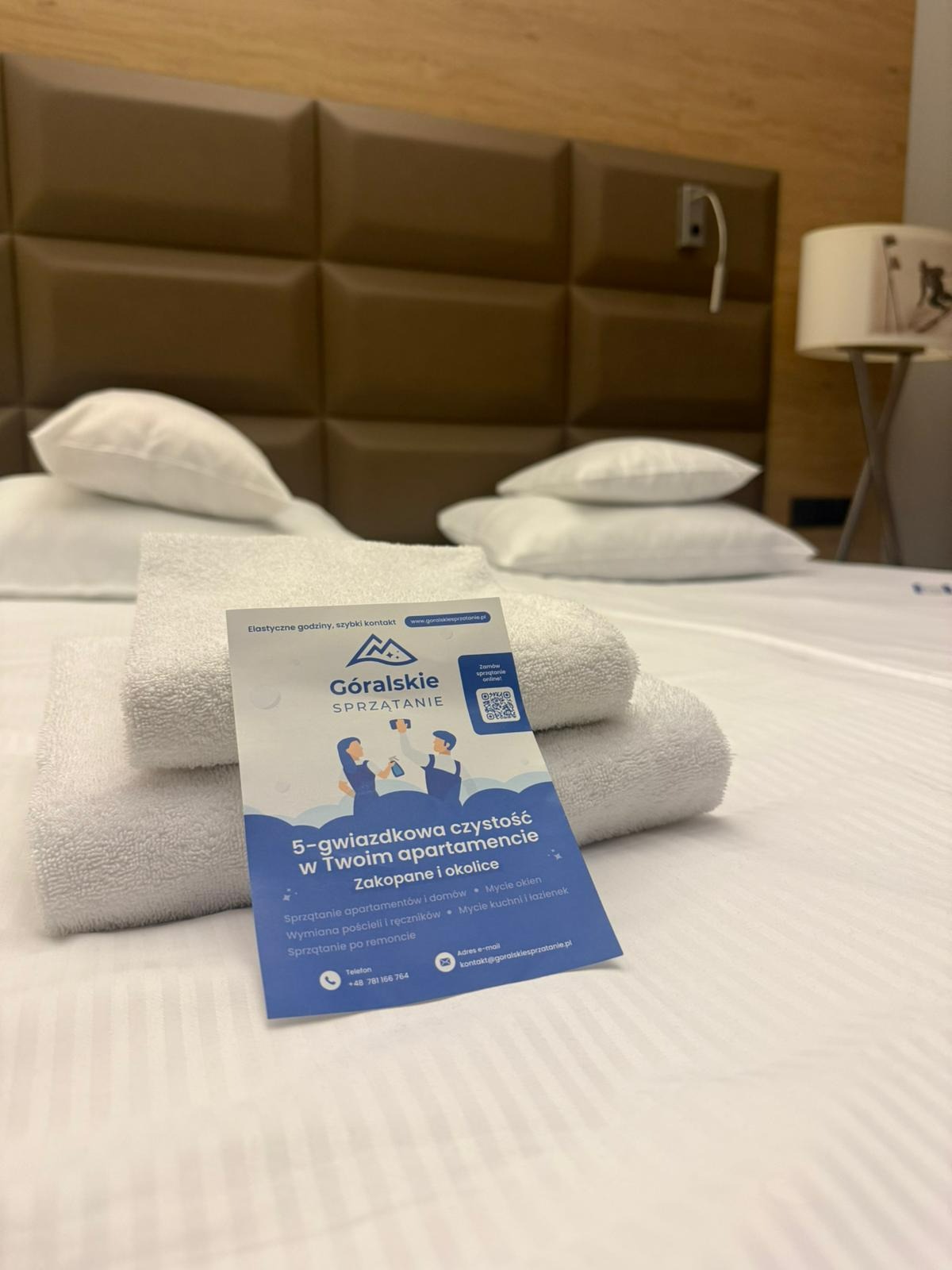 Stos białych ręczników i ulotka firmy sprzątającej apartamenty 'Góralskie Sprzątanie' na łóżku w pokoju hotelowym. Ulotka reklamuje sprzątanie 5-gwiazdkowe.