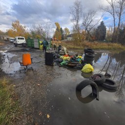 System A-Z - Opróżnianie terenu: pracownicy ładują odpady do kontenera, w tle ciężarówka i drzewa. Na pierwszym planie w wodzie opony i śmieci, obok pomarańczowe krzesło.