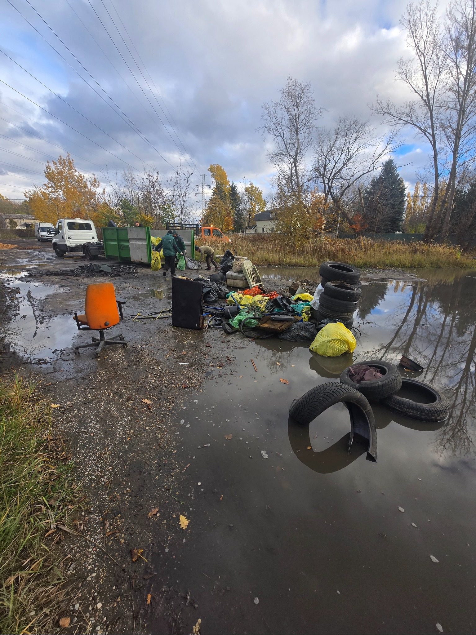 Opróżnianie terenu: pracownicy ładują odpady do kontenera, w tle ciężarówka i drzewa. Na pierwszym planie w wodzie opony i śmieci, obok pomarańczowe krzesło.