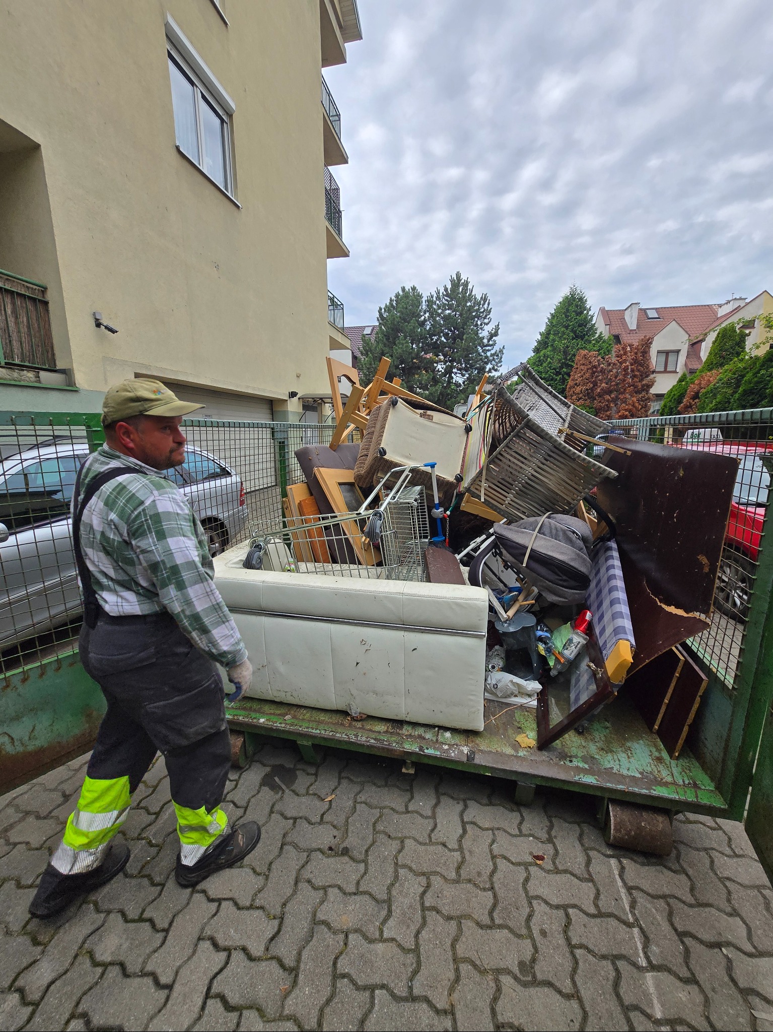 Pracownik w odblaskowych spodniach obok kontenera wypełnionego meblami i odpadami. W tle budynek mieszkalny i drzewa. Kontener na gruz i odpady.