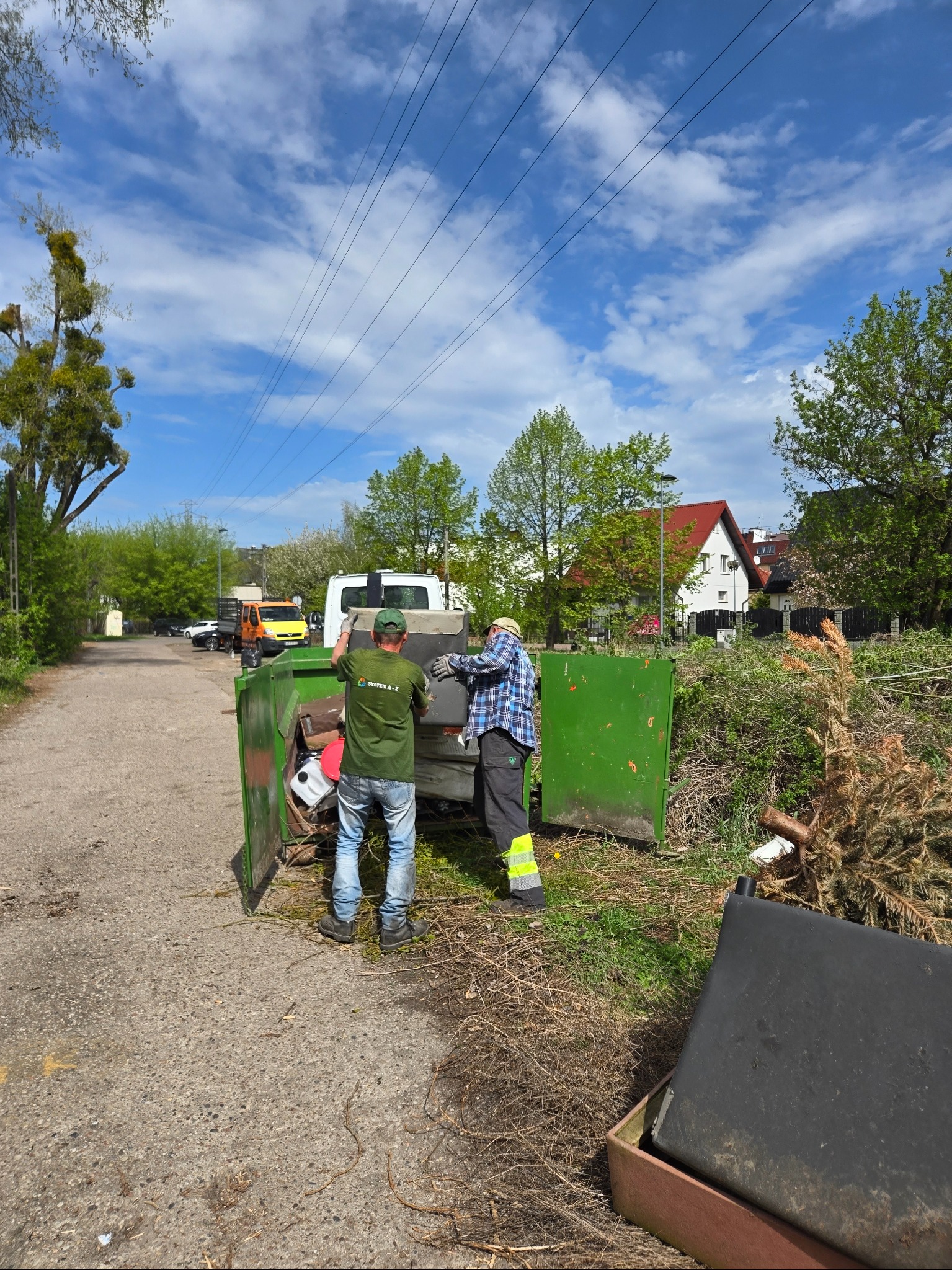 Dwóch pracowników wrzuca gałęzie i odpady do zielonego kontenera na przyczepie, na tle drzew i zabudowań. Widoczne niebo z chmurami.