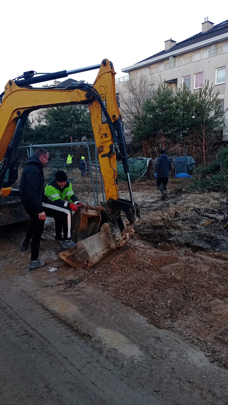 Żółta koparka z dwoma pracownikami w odblaskowych kamizelkach na placu budowy. W tle budynek mieszkalny i drzewa. Praca w toku, teren przygotowany pod inwestycję.