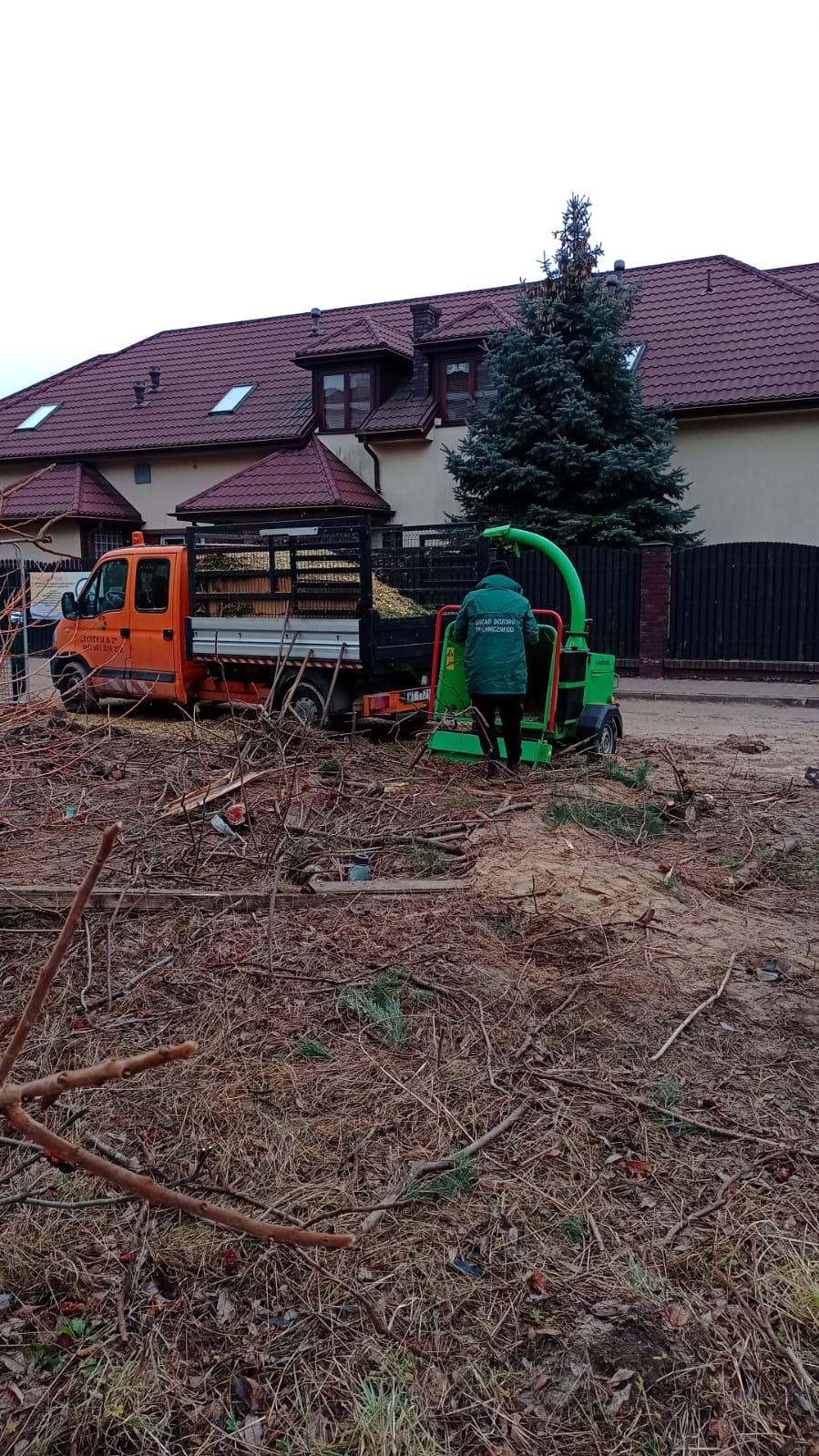 Pomarańczowy samochód z przyczepą wypełnioną gałęziami i zielony rozdrabniacz do gałęzi w trakcie pracy na tle domu z czerwoną dachówką i zielonego drzewa.