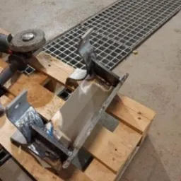 Element metalowy po spawaniu, szlifowany, leżący na palecie. W tle kratka ściekowa i narzędzia. Widoczne ślady obróbki i niedoskonałości materiału.