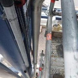 Aleksander Saja - Metalowa rura pionowa na rusztowaniu, mocowana pomarańczowymi opaskami. Widok z dołu, fragment buta w kadrze. Jasne tło, częściowo widoczne inne elementy konstrukcji.