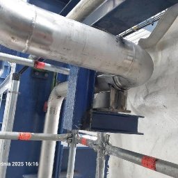 Aleksander Saja - Szczegółowy widok rurociągu ze stali nierdzewnej, wspartego na konstrukcji z niebieskich belek stalowych, z fragmentem rusztowania w dolnej części kadru.