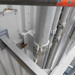 Metalowa konstrukcja wsporcza z rurami, mocowana do ściany, widoczna z dołu, z elementami rusztowania w tle. Detal techniczny.