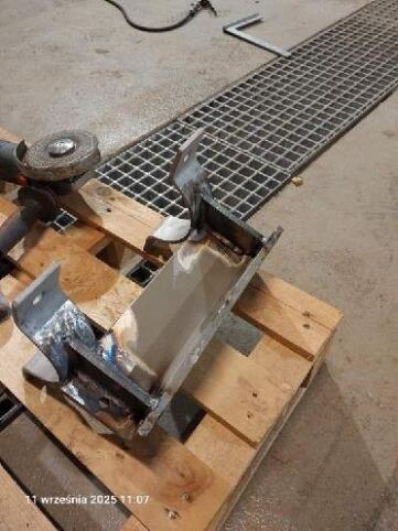 Metalowy element konstrukcyjny po spawaniu, leżący na palecie. Widoczny szlifierka kątowa i kratka ściekowa w tle. Detal z widocznymi śladami obróbki.