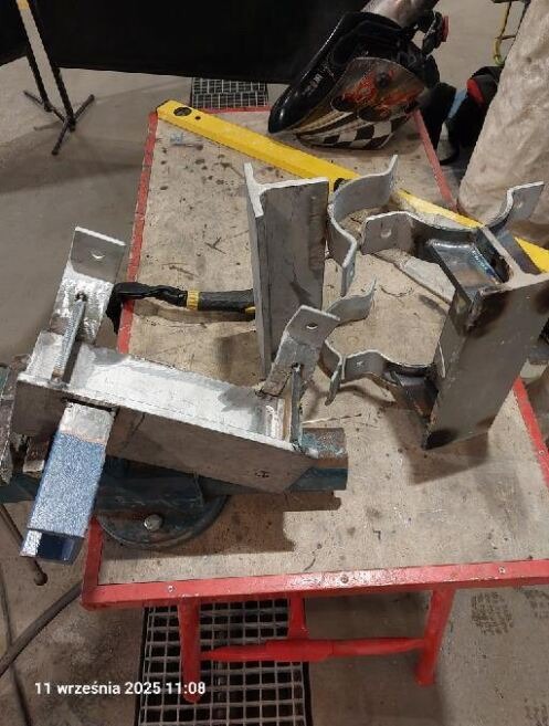 Metalowe elementy konstrukcyjne w trakcie obróbki ślusarskiej, uchwycone w kadrze z żółtą poziomicą i maską spawalniczą w tle na warsztatowym stole.