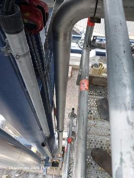 Metalowa rura pionowa na rusztowaniu, mocowana pomarańczowymi opaskami. Widok z dołu, fragment buta w kadrze. Jasne tło, częściowo widoczne inne elementy konstrukcji.