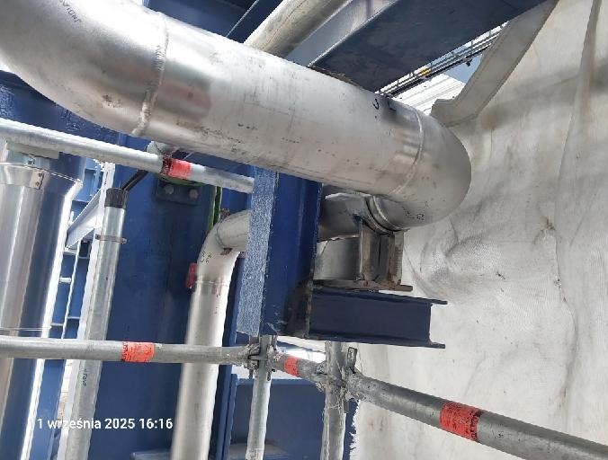 Szczegółowy widok rurociągu ze stali nierdzewnej, wspartego na konstrukcji z niebieskich belek stalowych, z fragmentem rusztowania w dolnej części kadru.