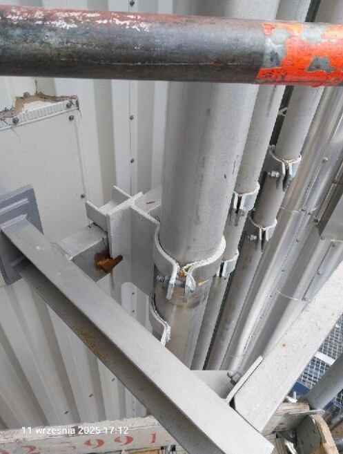 Metalowa konstrukcja wsporcza z rurami, mocowana do ściany, widoczna z dołu, z elementami rusztowania w tle. Detal techniczny.