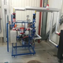 Złożona instalacja grzewcza z rurami w izolacji, manometrami i zaworami, zamontowana w pomieszczeniu o ścianach z blachy trapezowej. Widoczny fragment studzienki kanalizacyjnej.