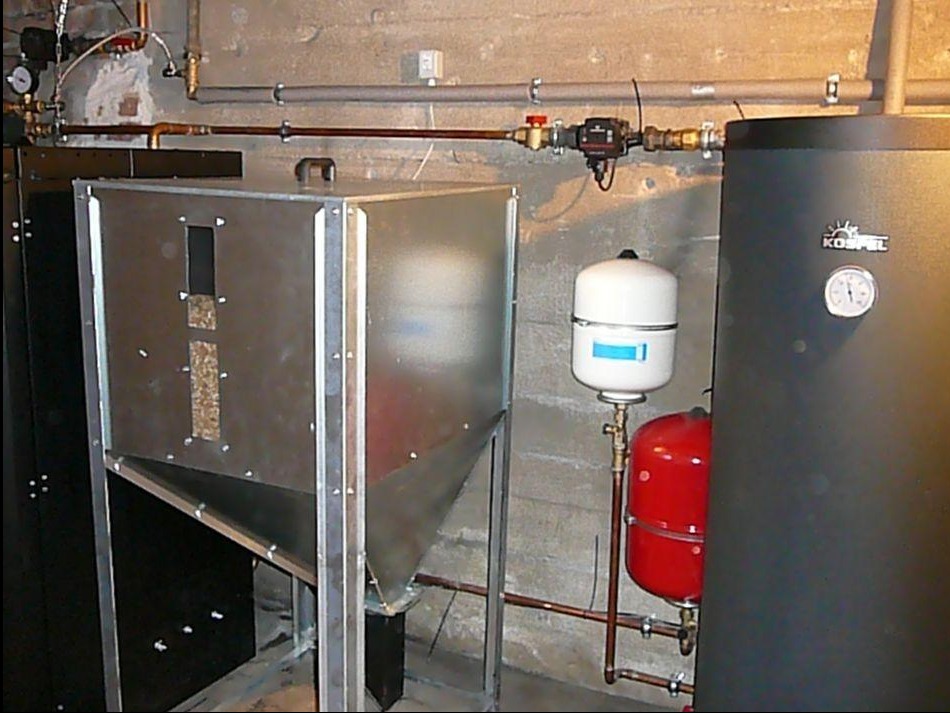 Instalacja kotła z podajnikiem, zbiornikiem i armaturą w kotłowni w Żukowie. Widoczne rury miedziane i elementy instalacji centralnego ogrzewania.