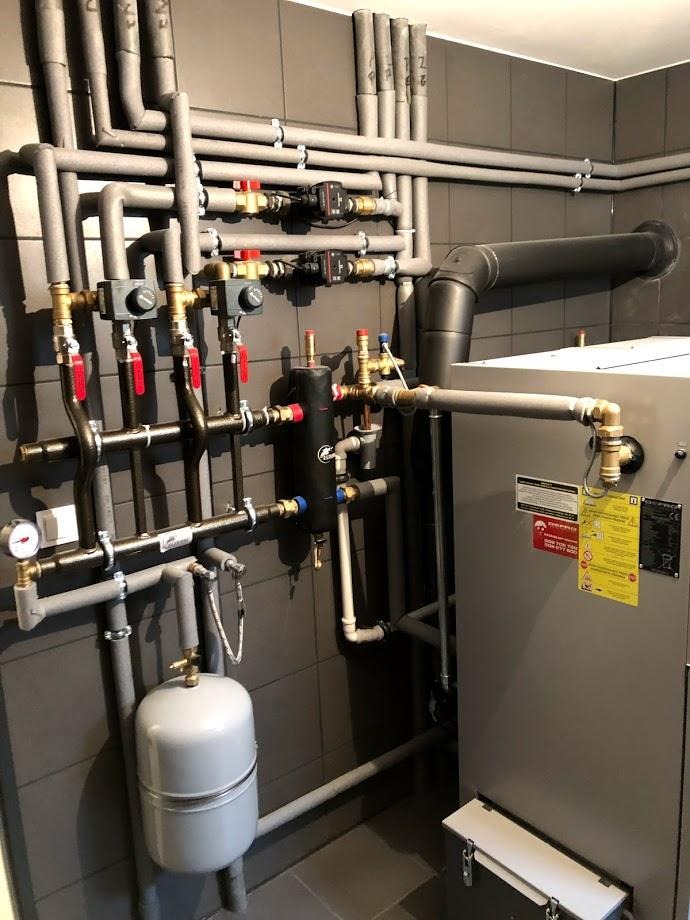 Złożona instalacja grzewcza z licznymi rurami, zaworami i zbiornikiem wyrównawczym na tle ciemnoszarych płytek ściennych. Widoczne elementy kotła i izolacja termiczna rur.