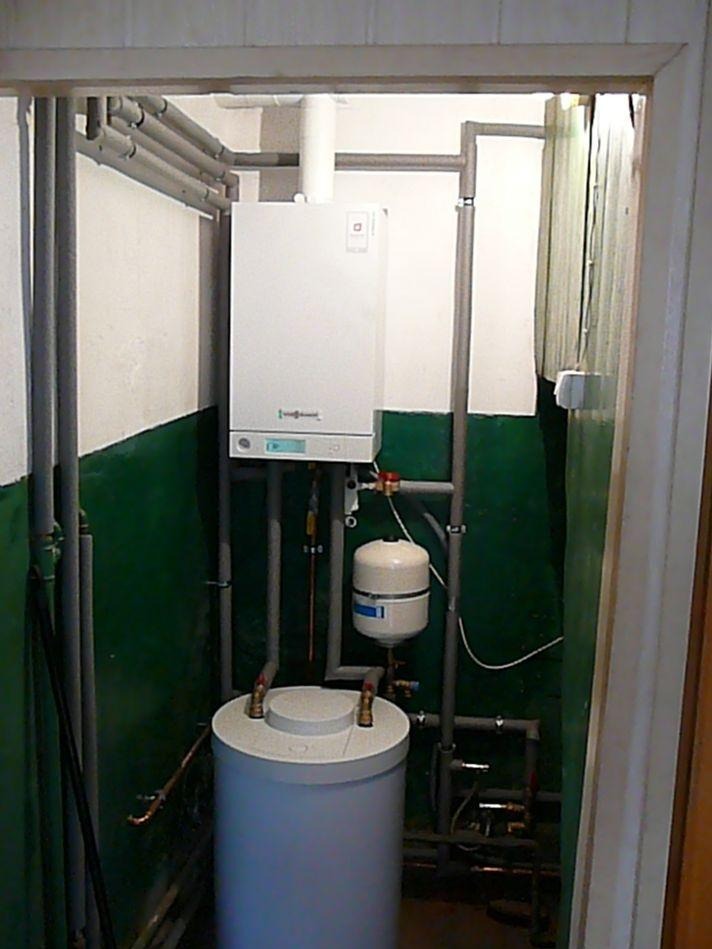 Biały piec gazowy Viessmann z instalacją CO, umieszczony w małym pomieszczeniu z zielonymi ścianami i szarymi rurami. Widok z bliska na zamontowany system.