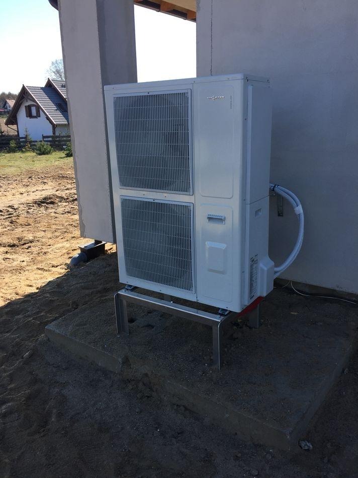 Biała pompa ciepła Viessmann na metalowym stelażu, zainstalowana na betonowej podstawie przy ścianie budynku w stanie surowym otoczona ziemią. Widoczny dom w tle.