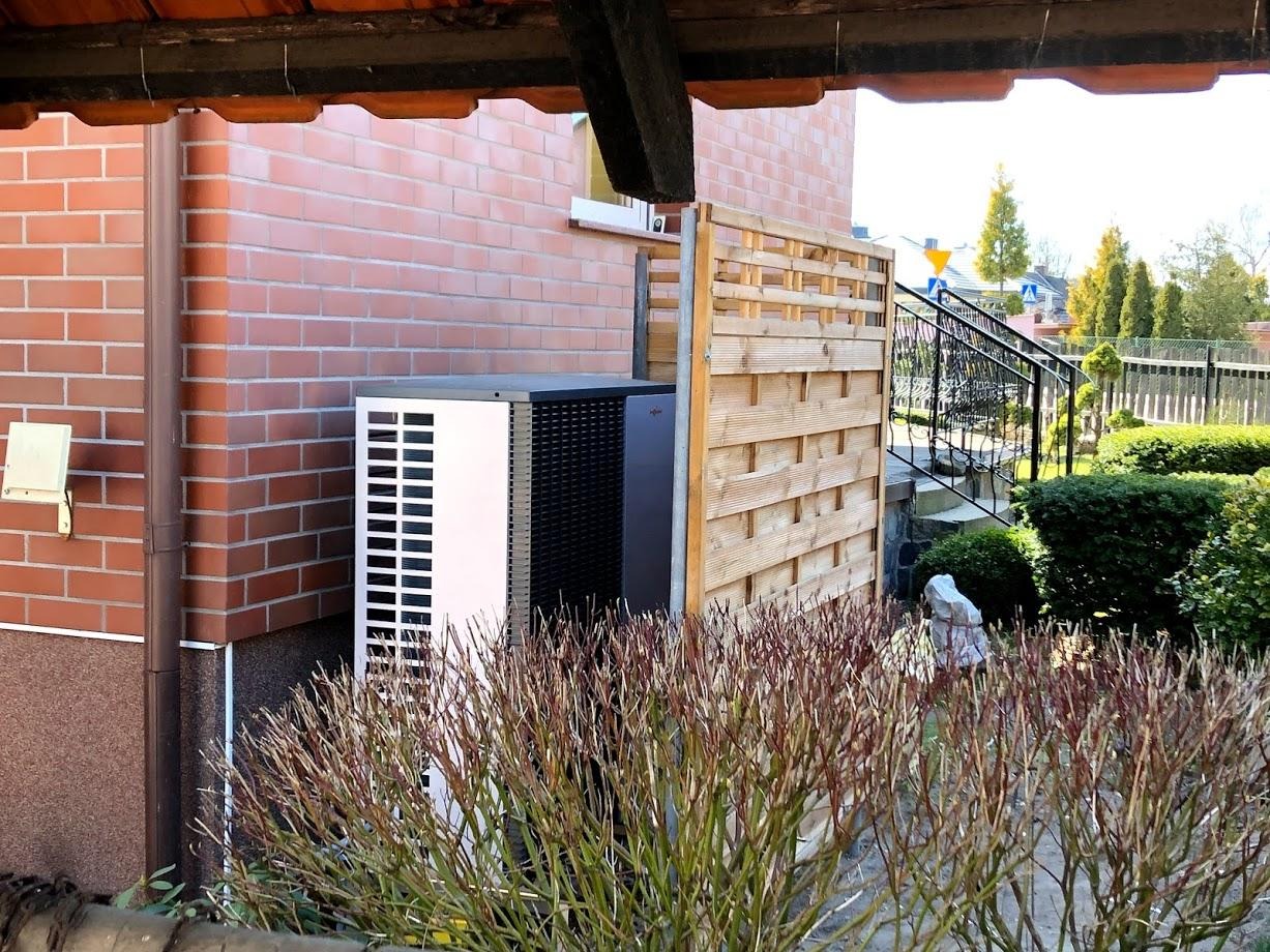 Biało-czarna pompa ciepła Viessmann na tle ceglanego budynku, częściowo zasłonięta drewnianym płotem. Widoczne krzewy na pierwszym planie i schody wejściowe do budynku.