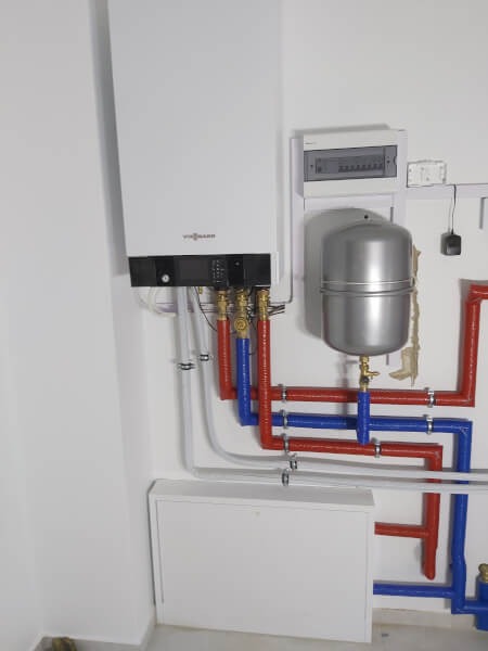 Biały piec gazowy Viessmann z podłączeniem rur w kolorach czerwonym i niebieskim na ścianie wewnątrz pomieszczenia. Widoczna instalacja i skrzynka elektryczna.