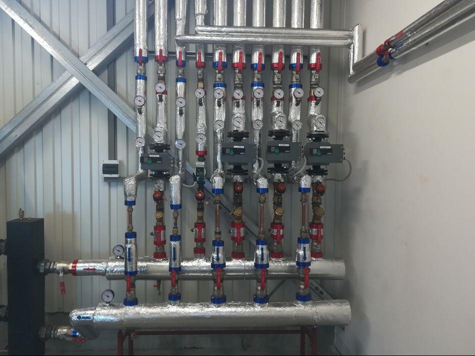 Szczegółowe ujęcie instalacji grzewczej z rurami w izolacji, zaworami i manometrami, zamontowanej przy ścianie z blachy trapezowej i białej ścianie.