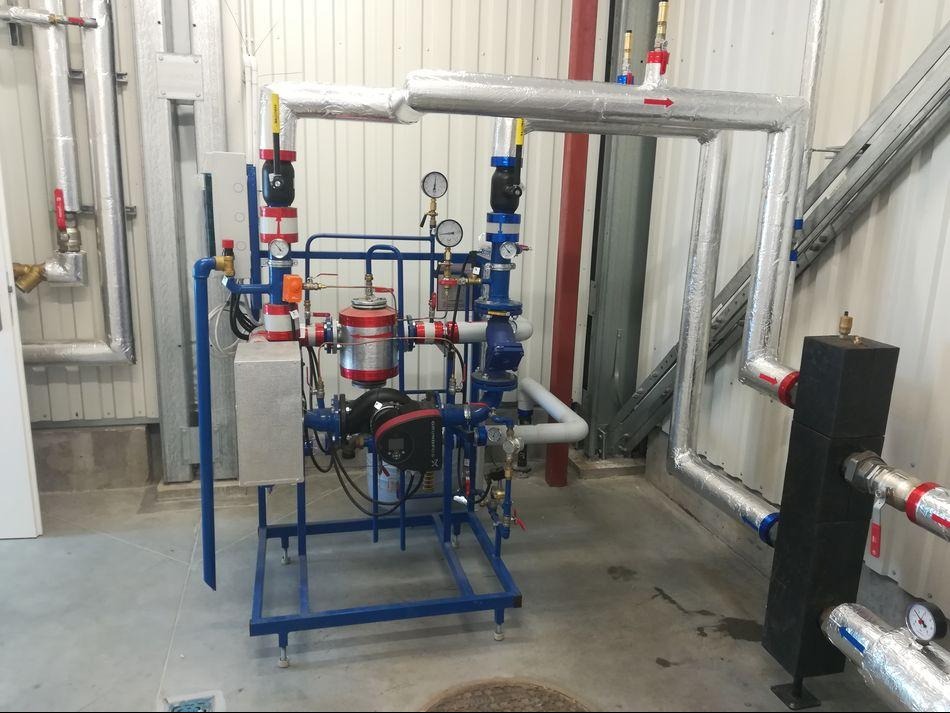 Złożona instalacja grzewcza z rurami w izolacji, manometrami i zaworami, zamontowana w pomieszczeniu o ścianach z blachy trapezowej. Widoczny fragment studzienki kanalizacyjnej.