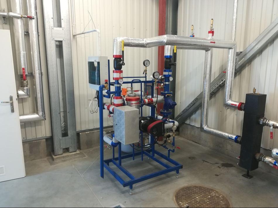 Złożona instalacja grzewcza z rurami w izolacji, manometrami i zaworami, umieszczona na mobilnej, niebieskiej ramie w hali przemysłowej. Widoczne elementy podłączenia do sieci.