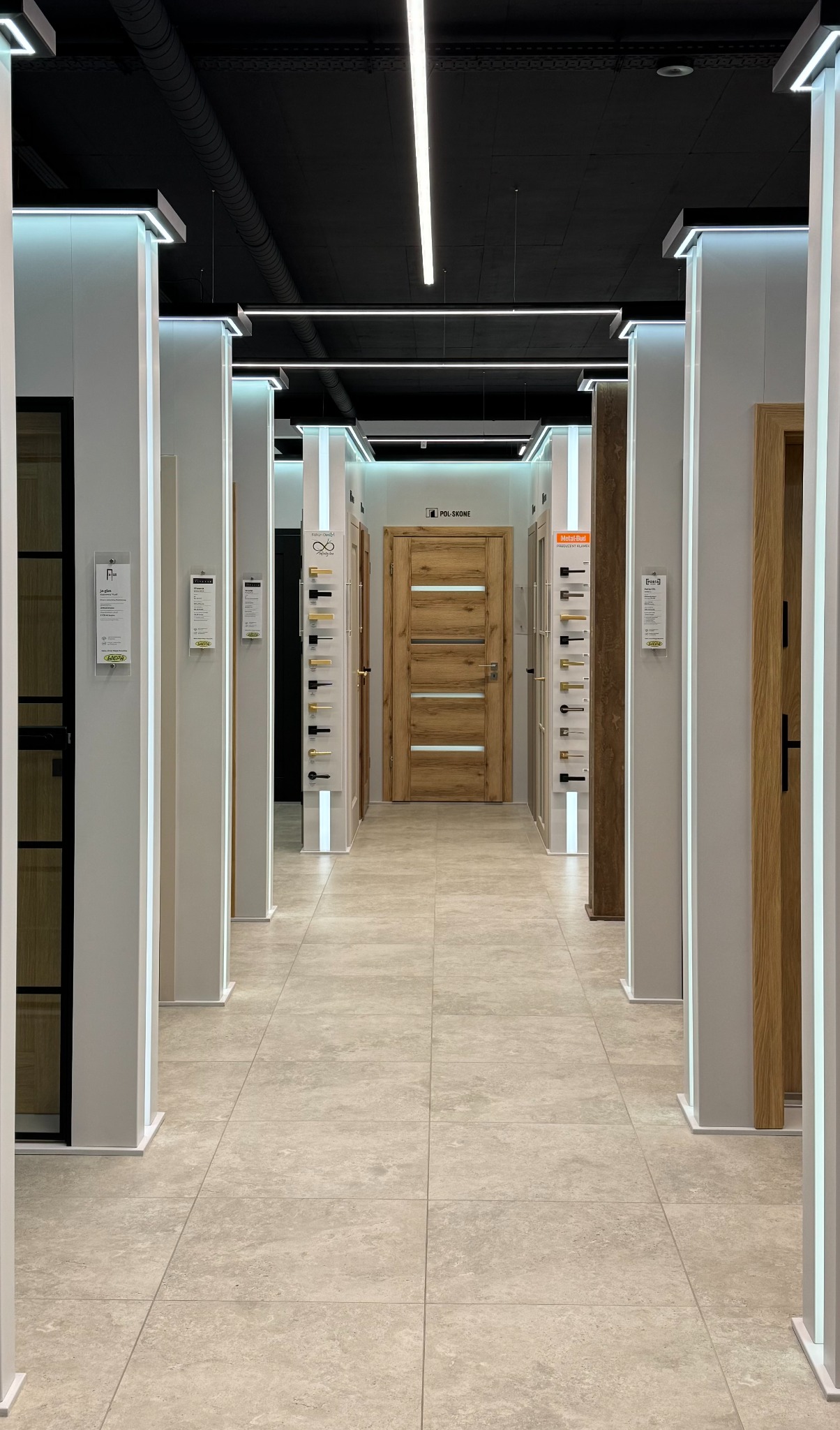 Salon z ekspozycją drzwi wewnętrznych różnych modeli, oświetlony ledami, z drewnianą okleiną i przeszkleniami, widok z perspektywy korytarza.