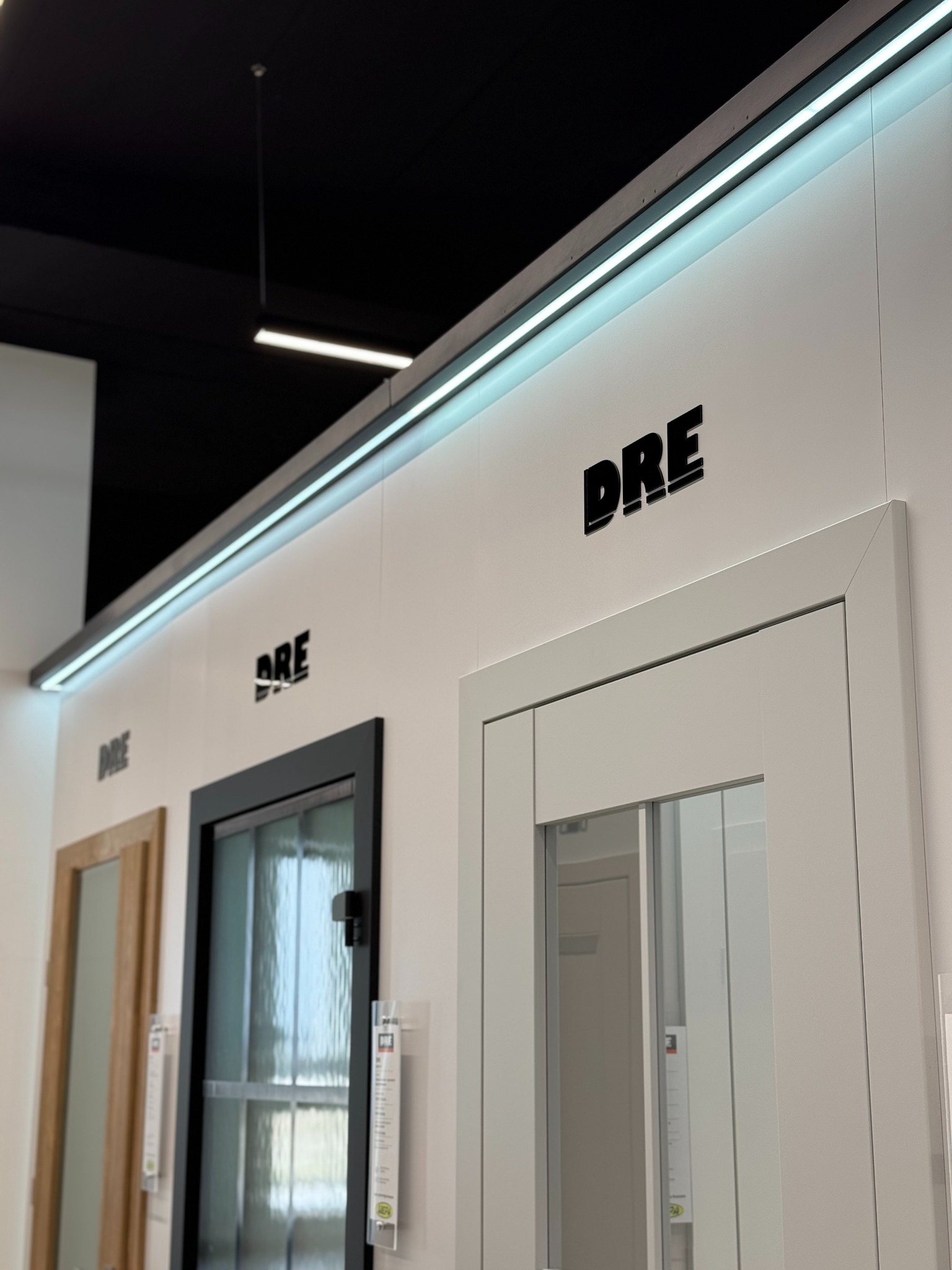 Ekspozycja modeli drzwi marki DRE w salonie: białe, czarne i drewniane, z oświetleniem LED. Widok z boku, z bliska na białe drzwi z przeszkleniem.