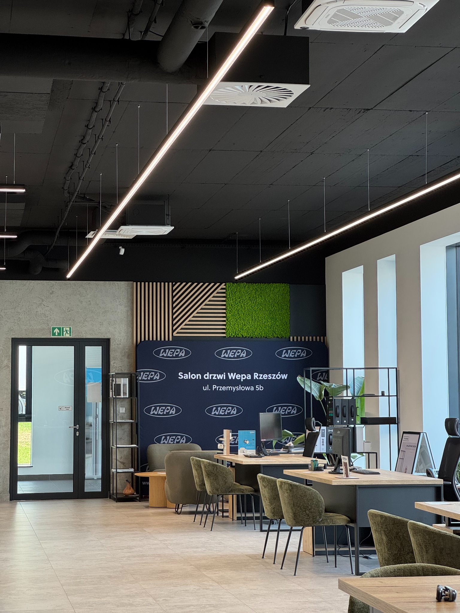 Wnętrze salonu drzwi Wepa Rzeszów z biurkami, krzesłami i ekspozycją drzwi. Widoczne logo firmy i adres na ścianie. Nowoczesny design z elementami zieleni.