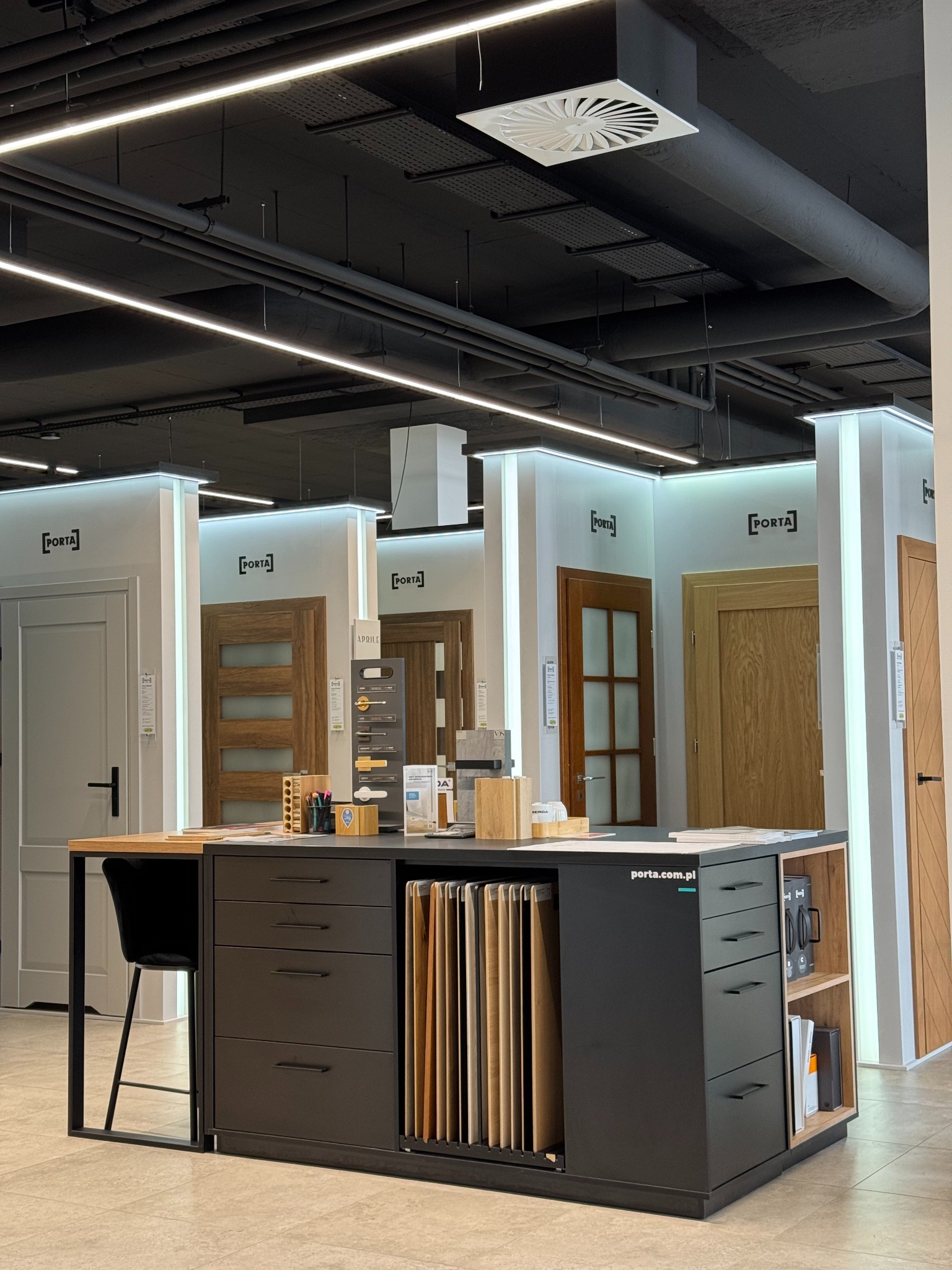 Ekspozycja modeli drzwi marki Porta w salonie sprzedaży. Widoczne próbki kolorów i materiałów wykończeniowych na blacie. Oświetlenie sufitowe i logo producenta.