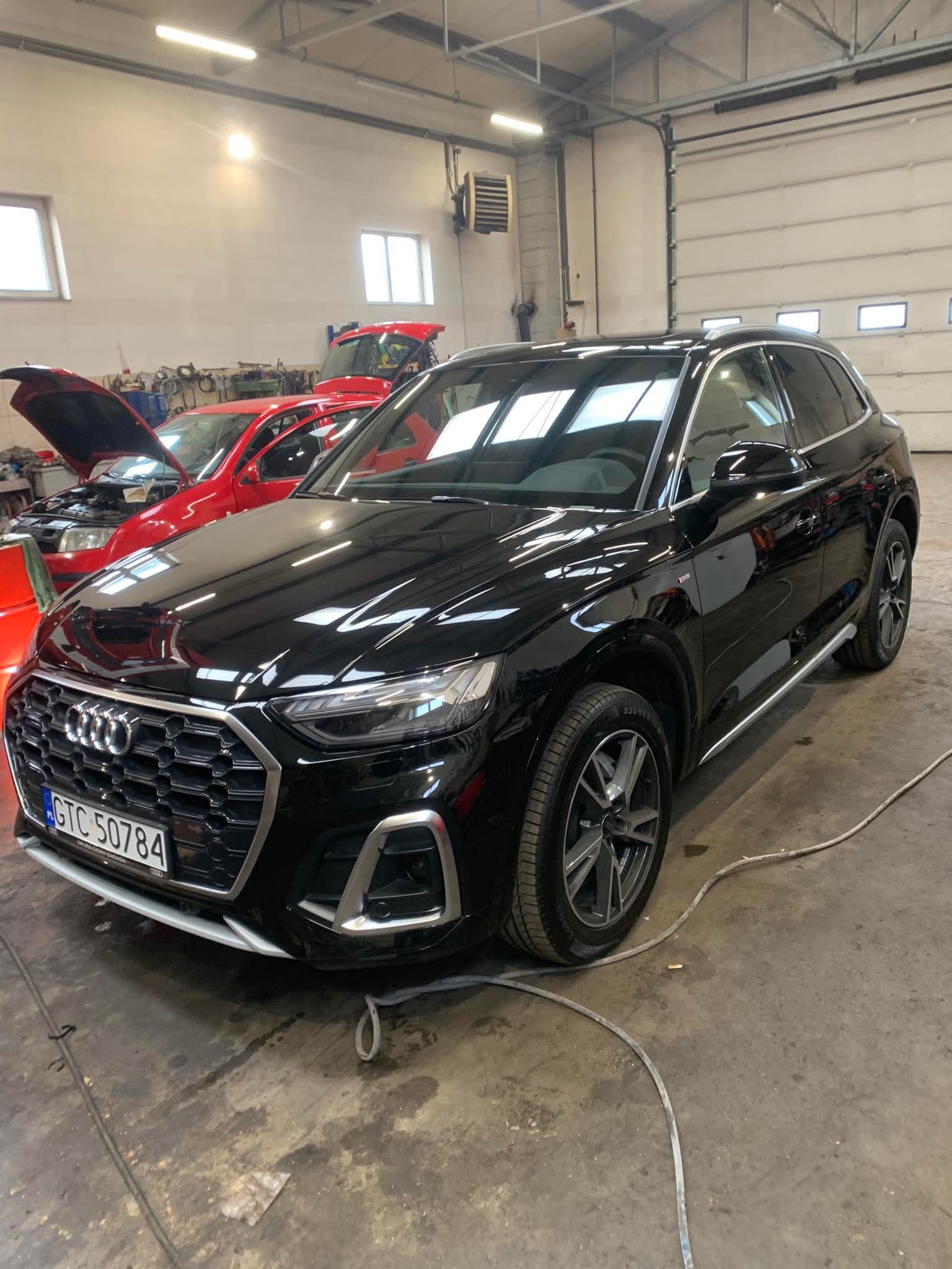 Lśniący, czarny SUV Audi Q5 w warsztacie, z widocznymi detalami karoserii i reflektorów. W tle czerwone auta z otwartymi maskami, sugerujące prace naprawcze.