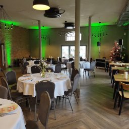 FABRYKA STYLU ILONA ŁYJAK - Sala bankietowa z okrągłymi stołami nakrytymi białymi obrusami, szarymi krzesłami, parkietem w jodełkę i zielonym oświetleniem, w tle choinka.