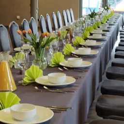 Długi stół z szarą tkaniną, nakryty białą zastawą, zielonymi serwetkami origami i kwiatami w wazonach. Krzesła z szarym obiciem wzdłuż stołu.