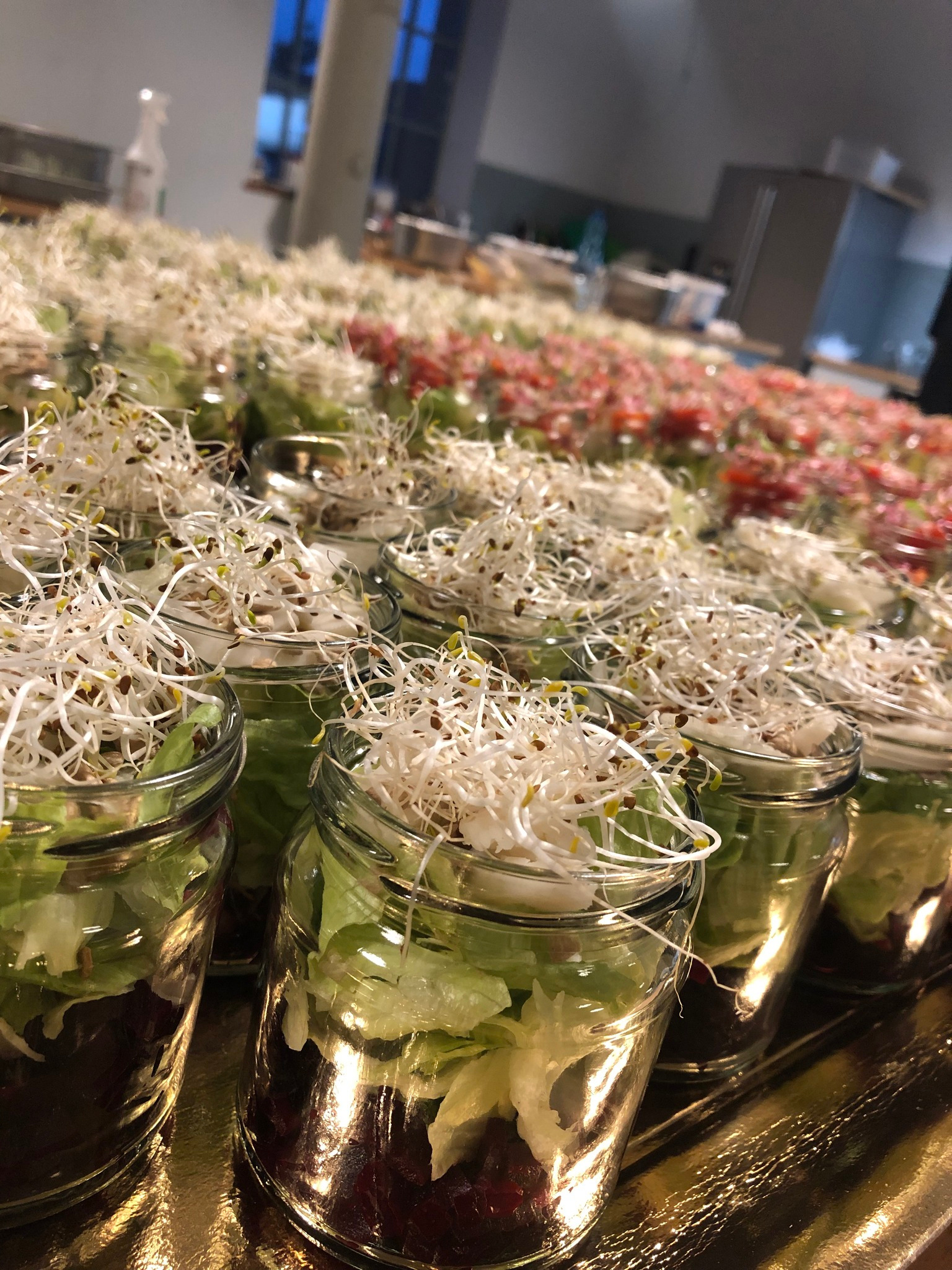 Szereg słoików z sałatkami warstwowymi, udekorowanymi kiełkami, ustawionych na złotej tacy. W tle widoczne kolejne porcje, przygotowane do serwowania.