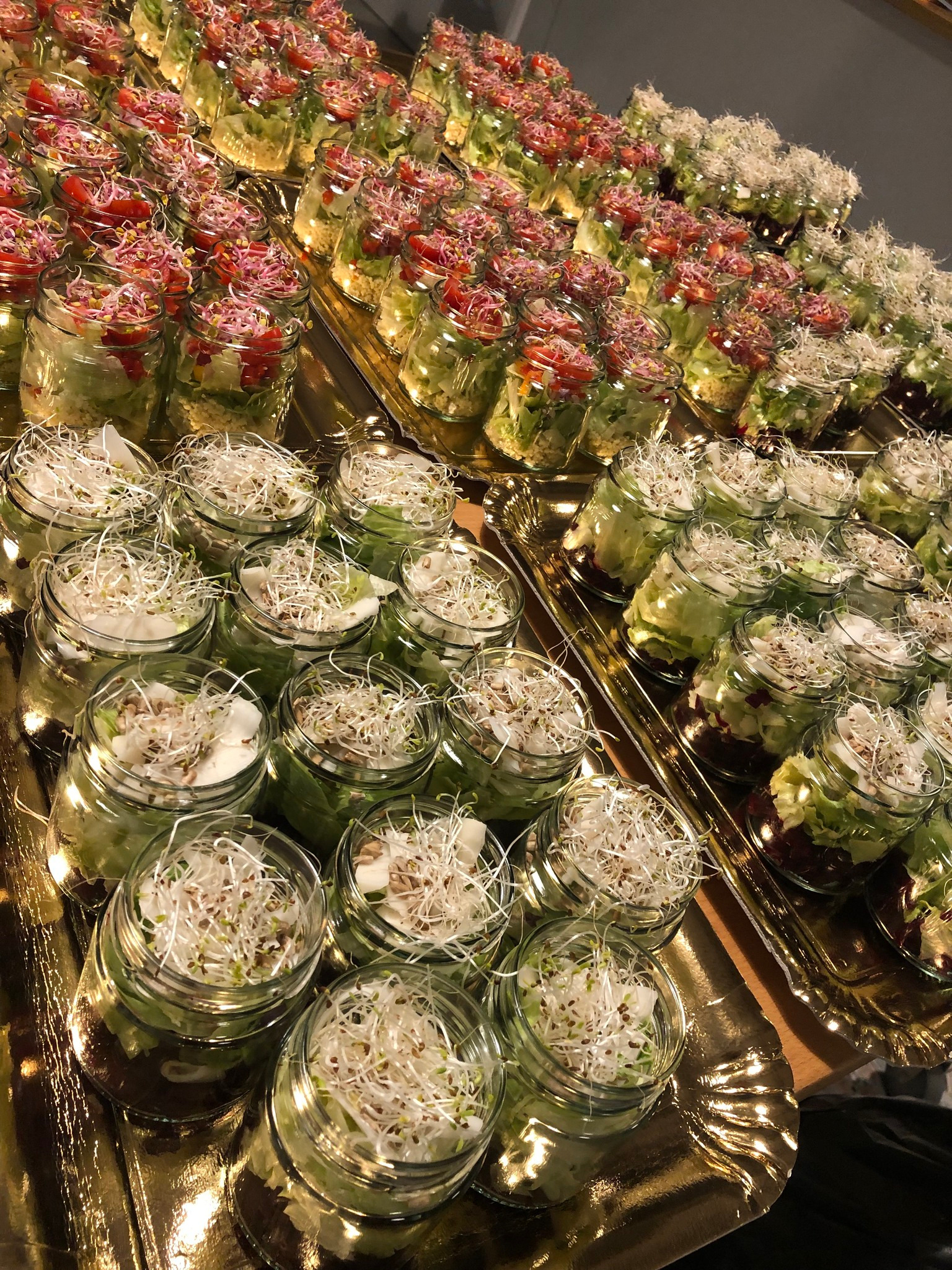 Szklane słoiki z sałatkami na złotych tacach. Różne kompozycje: z kiełkami, pomidorami i czerwoną kapustą. Catering na imprezę, detale.