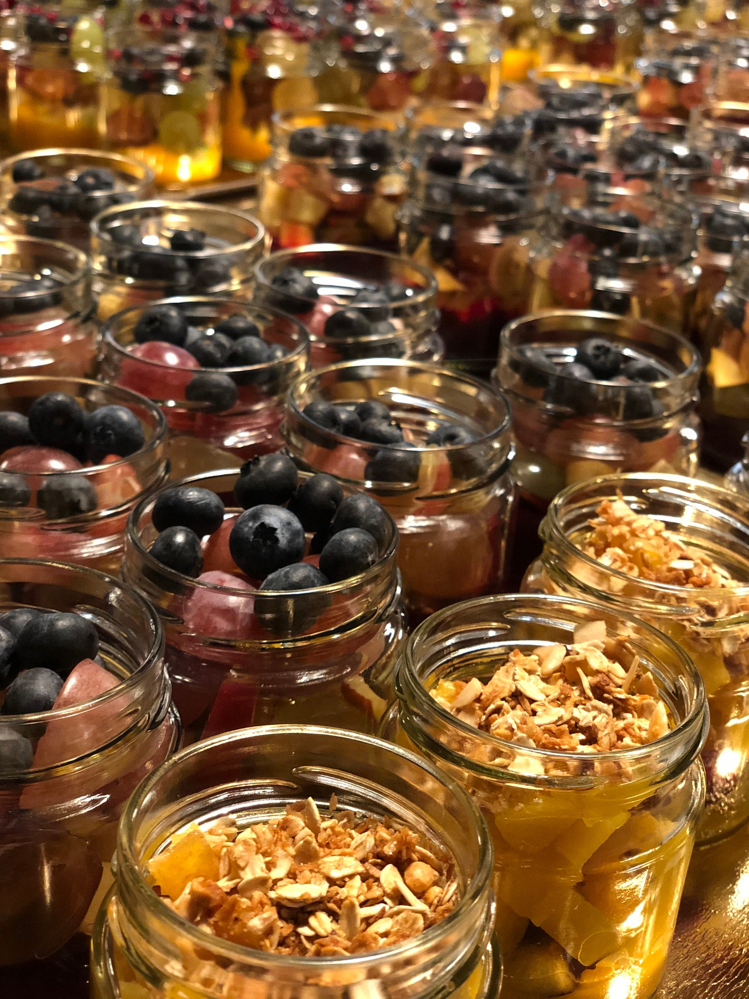 Szereg słoiczków z owocami i granolą, gotowych do podania. Różne kompozycje kolorystyczne i smaki, idealne na catering imprezowy. Widok z bliska.