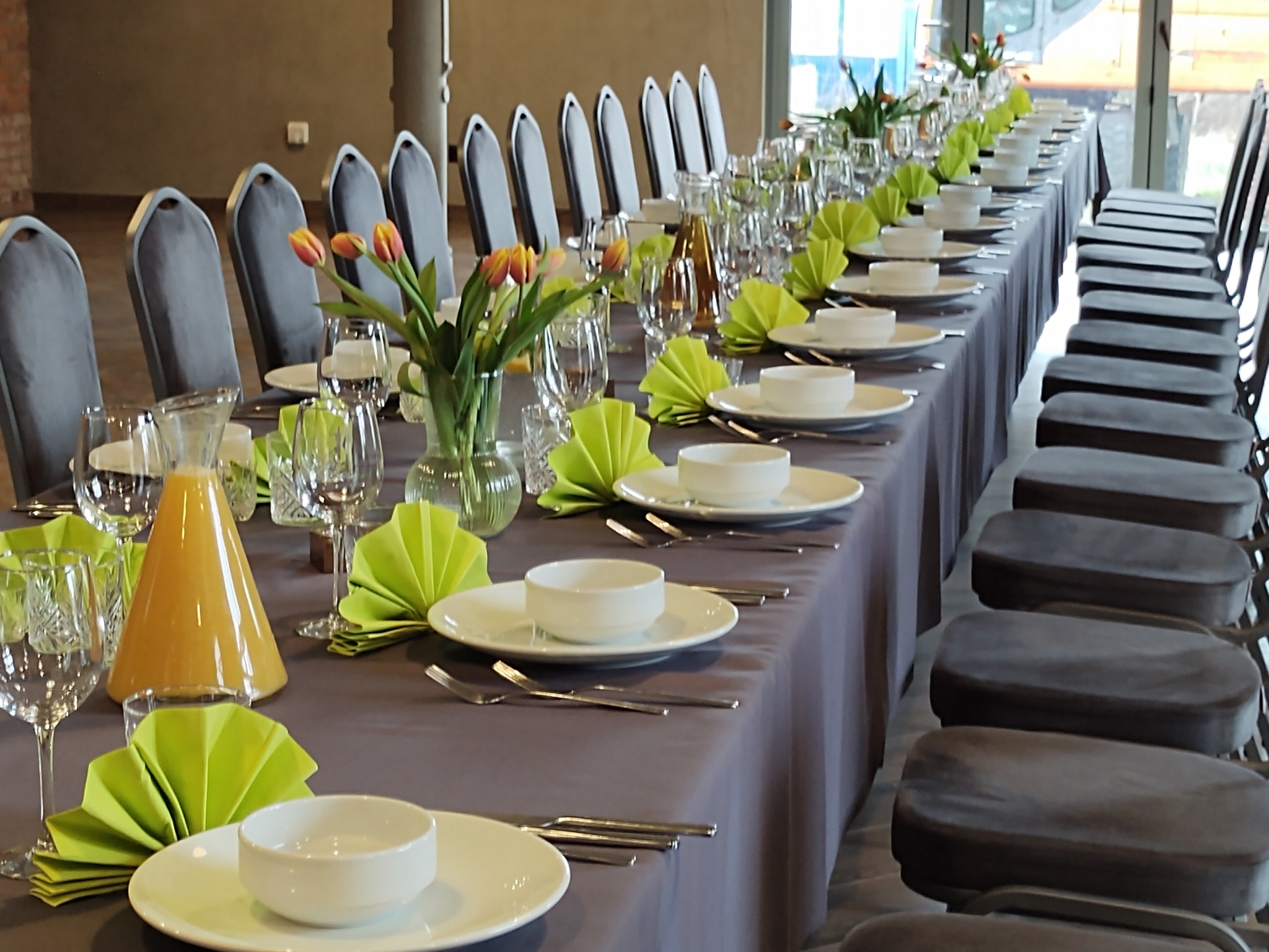 Długi stół z szarą tkaniną, nakryty białą zastawą, zielonymi serwetkami origami i kwiatami w wazonach. Krzesła z szarym obiciem wzdłuż stołu.