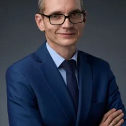 Portret mężczyzny w okularach i niebieskim garniturze, z założonymi rękami, na szarym tle. Uśmiechnięty, z zegarkiem na ręku i obrączką na palcu.