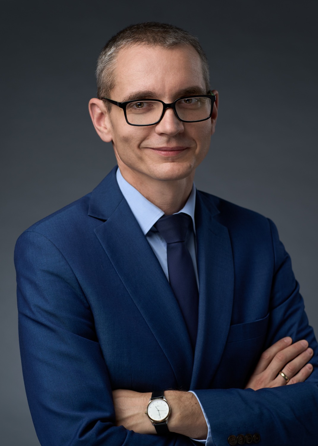 Portret mężczyzny w okularach i niebieskim garniturze, z założonymi rękami, na szarym tle. Uśmiechnięty, z zegarkiem na ręku i obrączką na palcu.