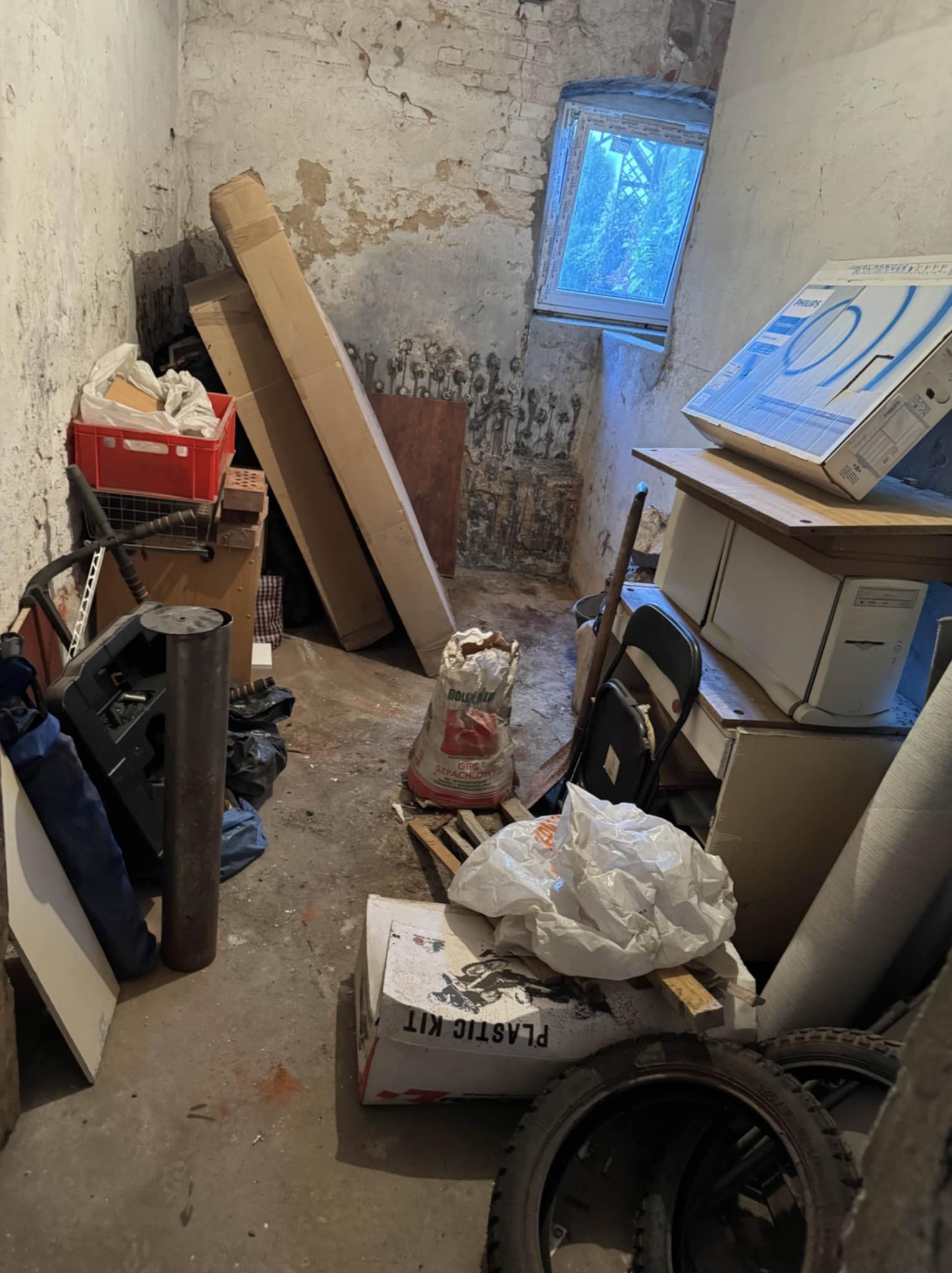 Zaniedbane pomieszczenie z oknem, pełne kartonów, worków, opon i innych przedmiotów. Widoczne ślady wilgoci na ścianach i podłodze. Niewielkie okno w tle doświetla wnętrze.