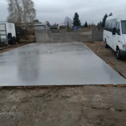 Wylana, gładka, betonowa płyta fundamentowa na działce, z widocznym szalunkiem, obok zaparkowany biały bus, w tle ogrodzenie z prefabrykatów betonowych i drzewa.