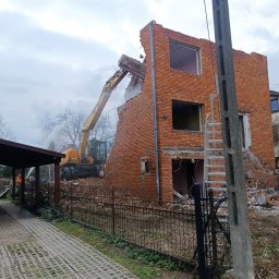 TRELBUD Klaudia Trela - Żółta koparka z hydraulicznym młotem w trakcie rozbiórki ceglanego budynku. Widoczny fragment chodnika z betonowych płyt i słup elektryczny.