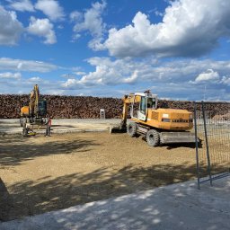 TRELBUD Klaudia Trela - Utwardzanie terenu: Żółta koparka Liebherr wyrównuje ziemię, w tle stos drewna i błękitne niebo z chmurami. Pracownik zagęszcza podłoże ubijarką.